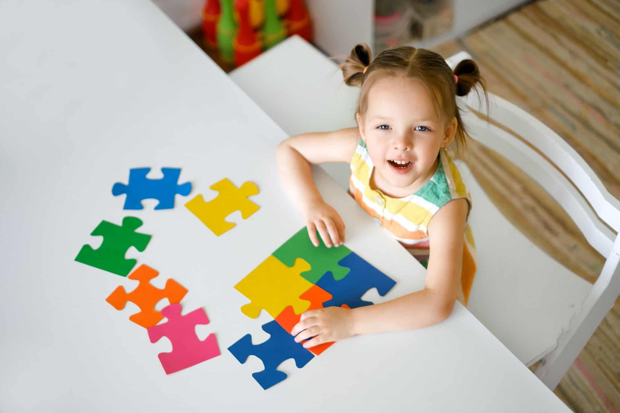 Girl is sitting at a table and assembling a puzzle of elements on autism Day.
